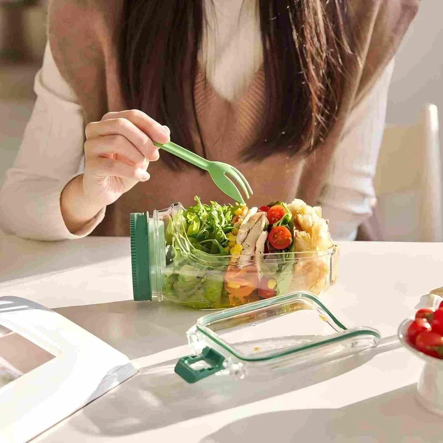 Person eating a salad from a portable container with a fork, sitting at a table.